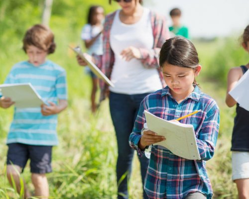 An elementary school girl of Filipino ethnicity is outdoors with her class exploring nature. She is busy writing on her notepad while walking with the group.