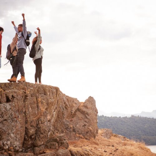 A group of five happy young adult friends cheer with their arms in the air at the summit of a mountain during a hike