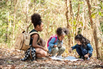 Group of Diversity little girl friends with backpack hiking together at forest mountain in summer sunny day. Three kids having fun outdoor activity sitting and looking at the map exploring the forest.