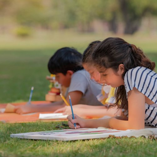 Two young girls and a boy are drawing a picture while lying down in the park.